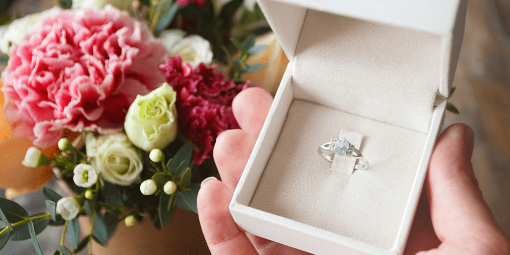 Person Holding Restored Aquamarine Gemstone Ring Displayed in Box Next to Bouquet of Flowers